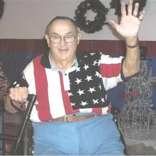 Picture of Robert Heft sitting on a couch wearing a shirt with the modern American Flag on it, the flag he designed at 17 years old. Source: https://www.wakemanfuneralhome.com/local-info-and-resources/bob-heft-and-the-50-star-flag